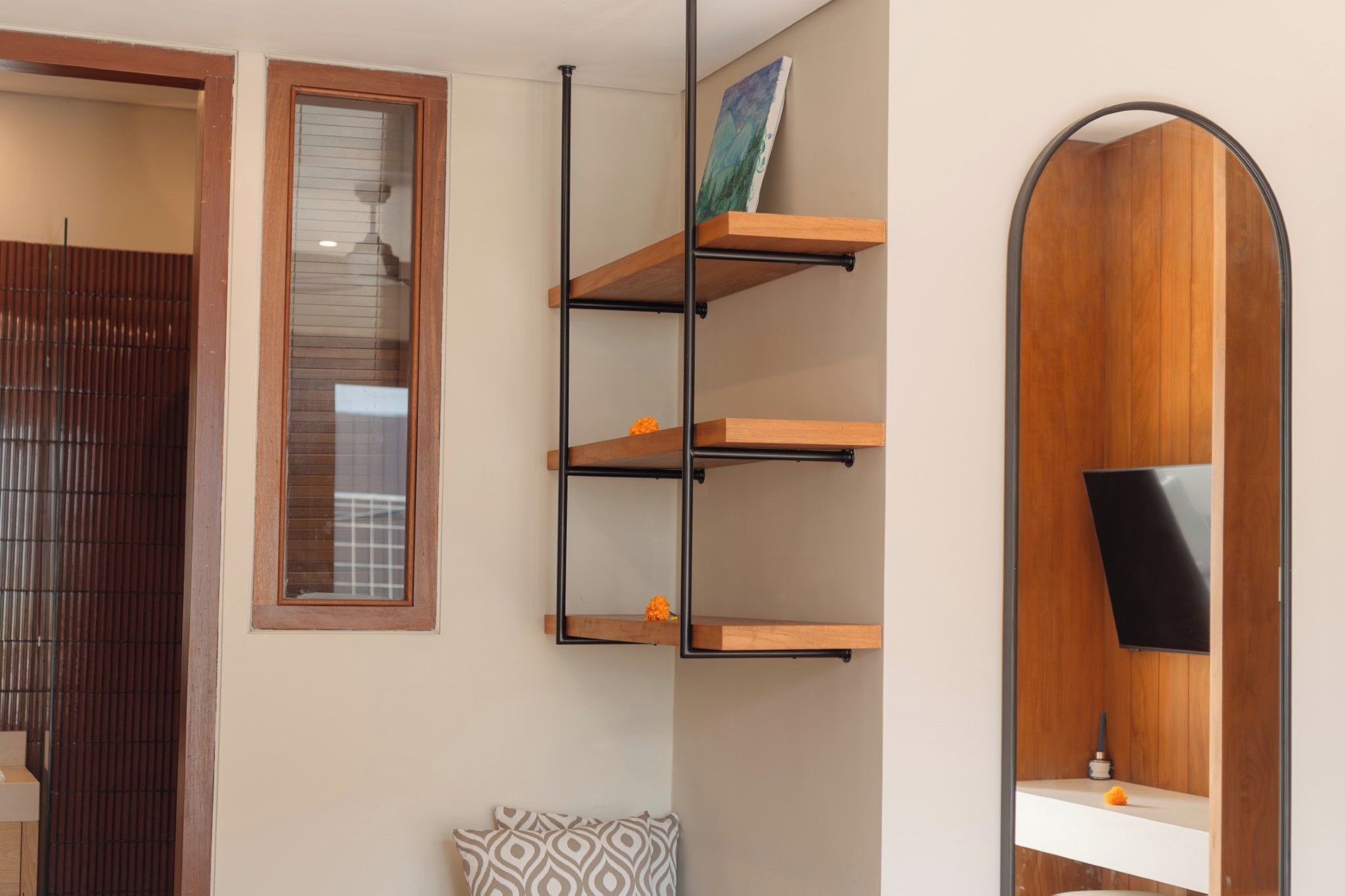 A room with a wooden door, a window with blinds, a wall-mounted shelf with a painting, a mirror, and a television.