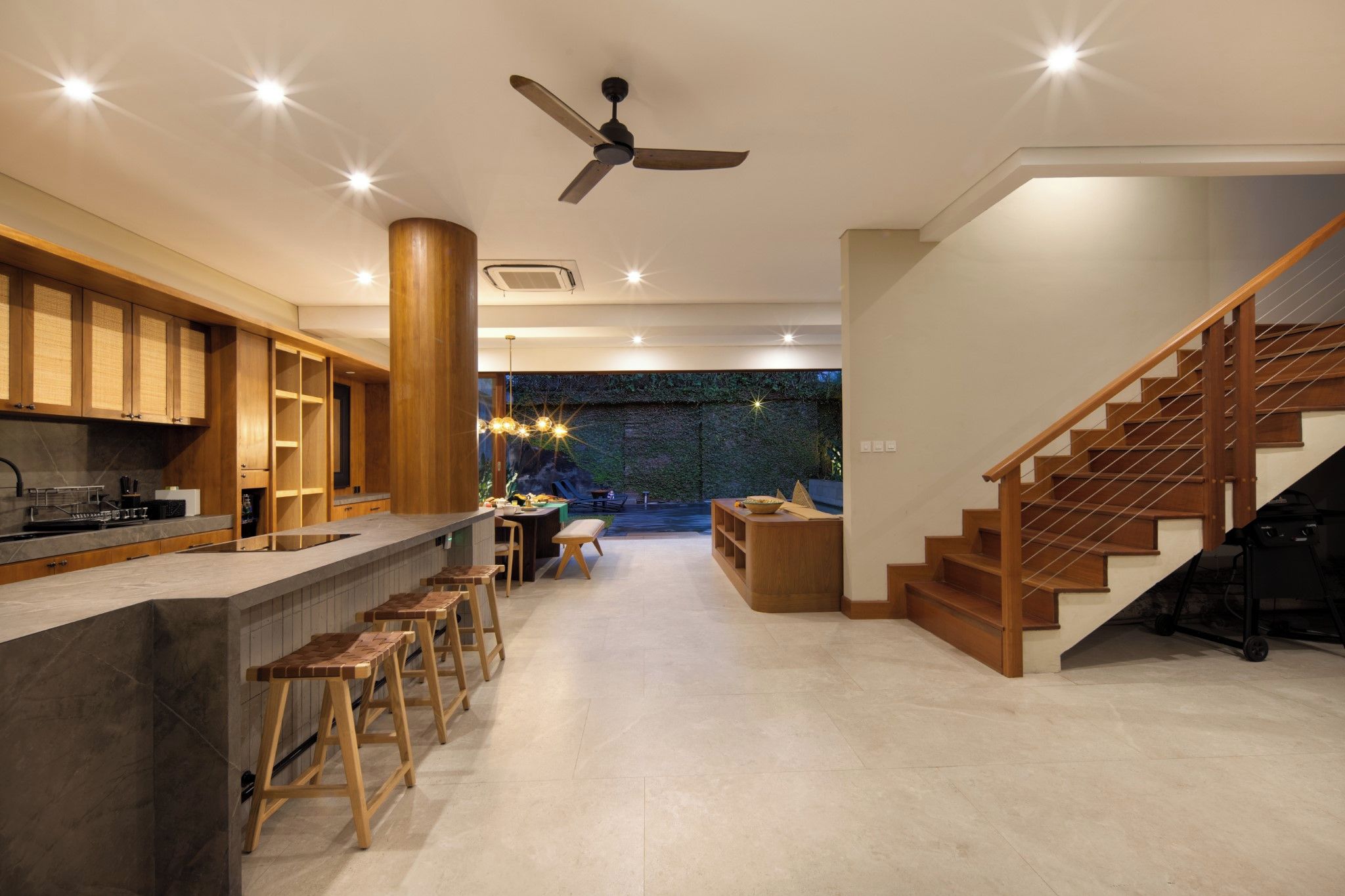 A modern kitchen with a bar area, wooden stools, and a staircase leading to a living area with a view of a green wall.