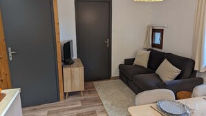 A compact living space with a dark door, a small wooden side table with a TV, a dark sofa with cushions, a dining table with a plate and wine glass, and a hanging light fixture. - Gîte La P'tite Lulu à St Pierre D'entremont, à Côté Cirque de St Même (Saint-Pierre-d'Entremont)