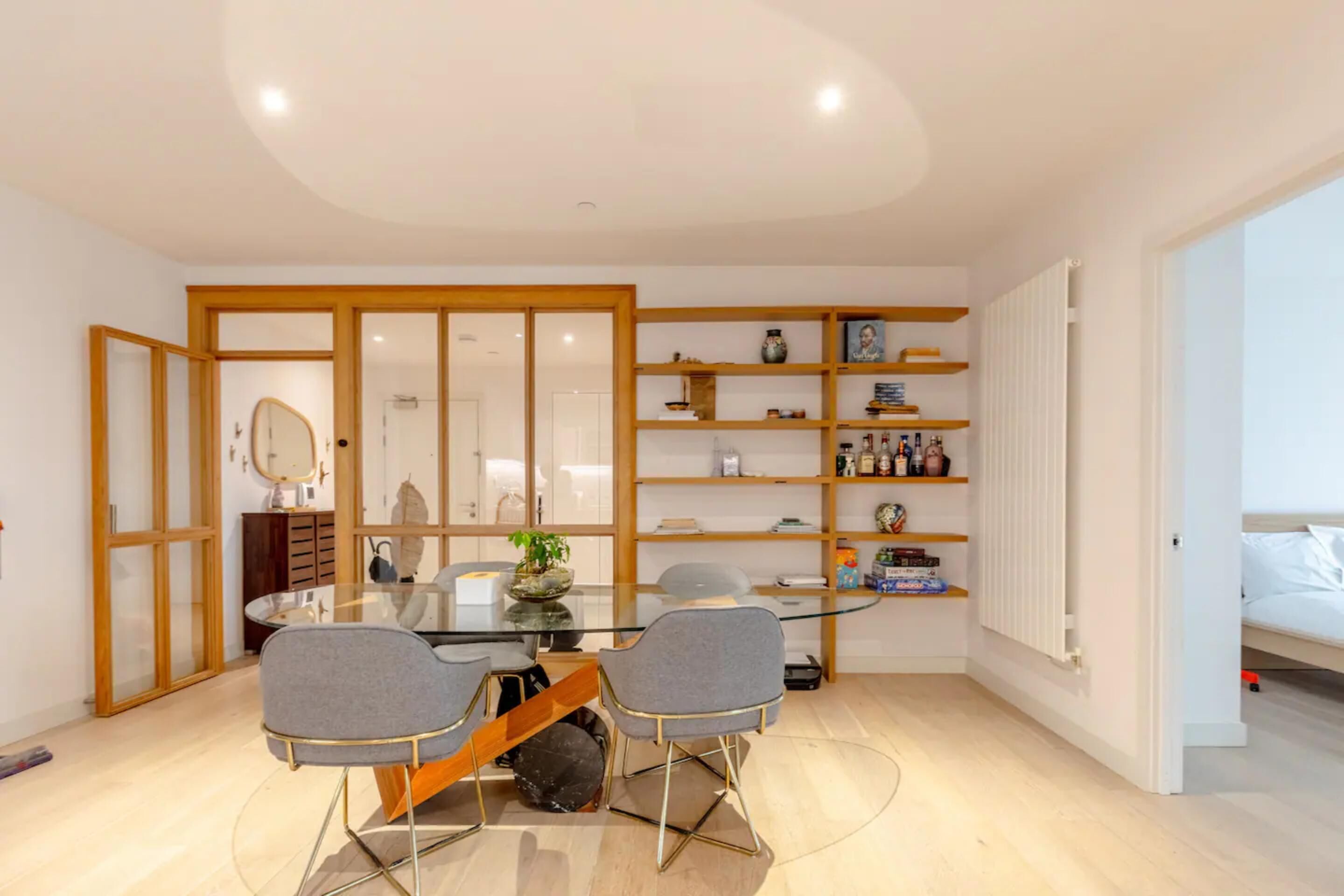 A modern dining area with a glass table, two grey chairs, wooden shelves with books and decorative items, and a white radiator.