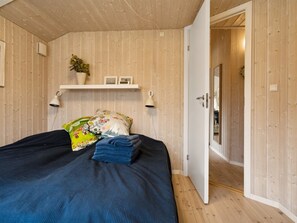 A bedroom with a wooden ceiling, a bed with a blue bedspread, a floral pillow, and a small shelf with decorative items.