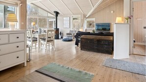 Een ruime woonkamer met een hoge nok, houten balken, een open haard, een eethoek en een zithoek met een televisie.