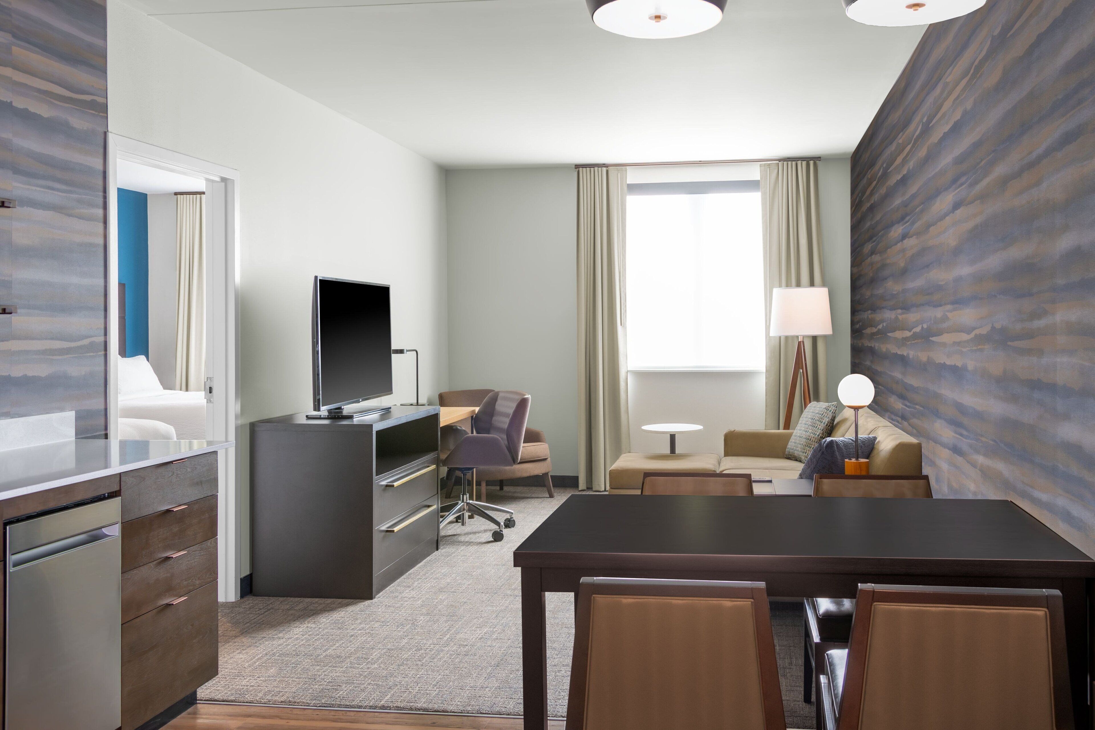 A hotel room with a desk, chair, TV, sofa, and a dining table with chairs.