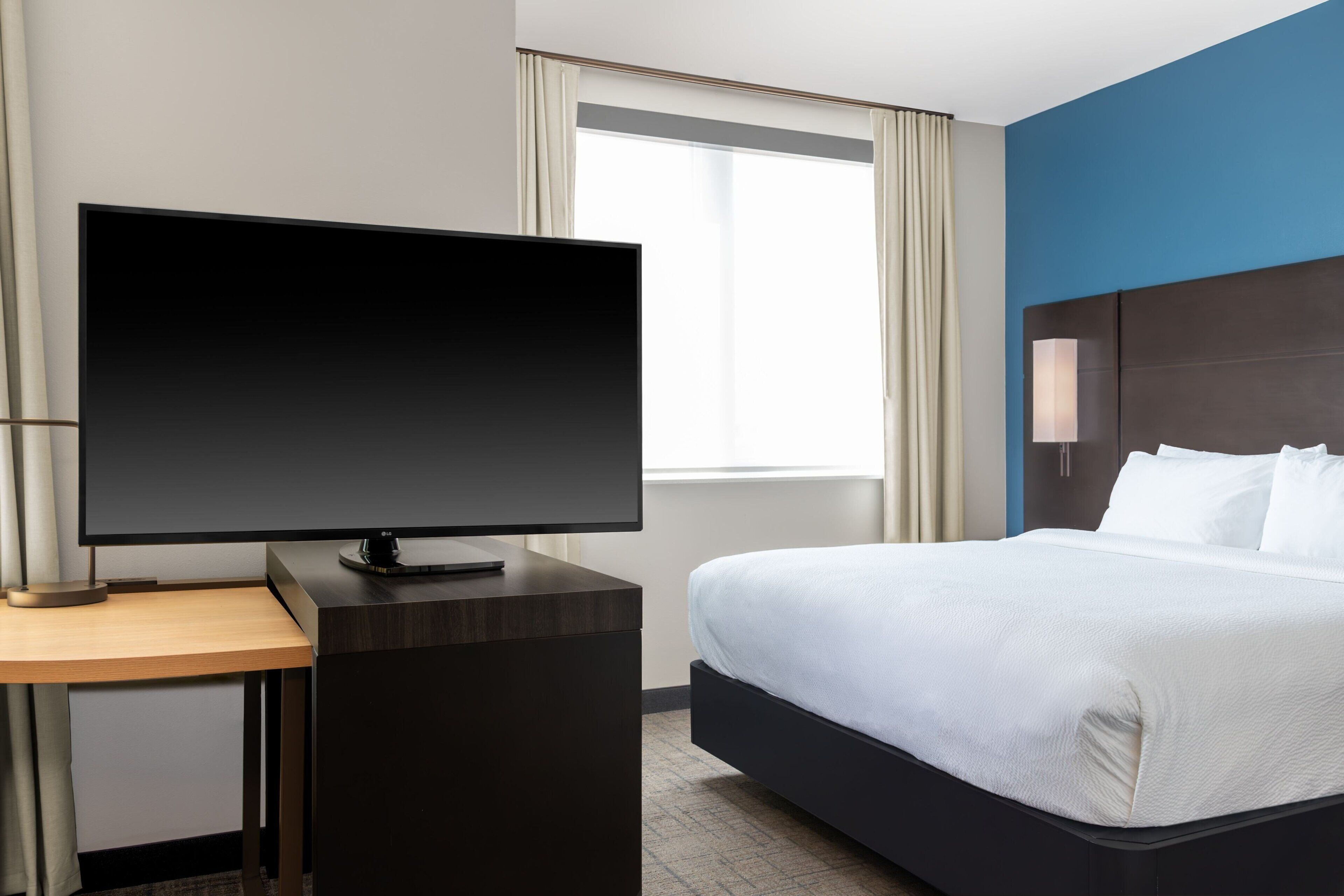 A hotel room with a bed, a desk with a flat-screen TV, a window with curtains, and a nightstand with a lamp.