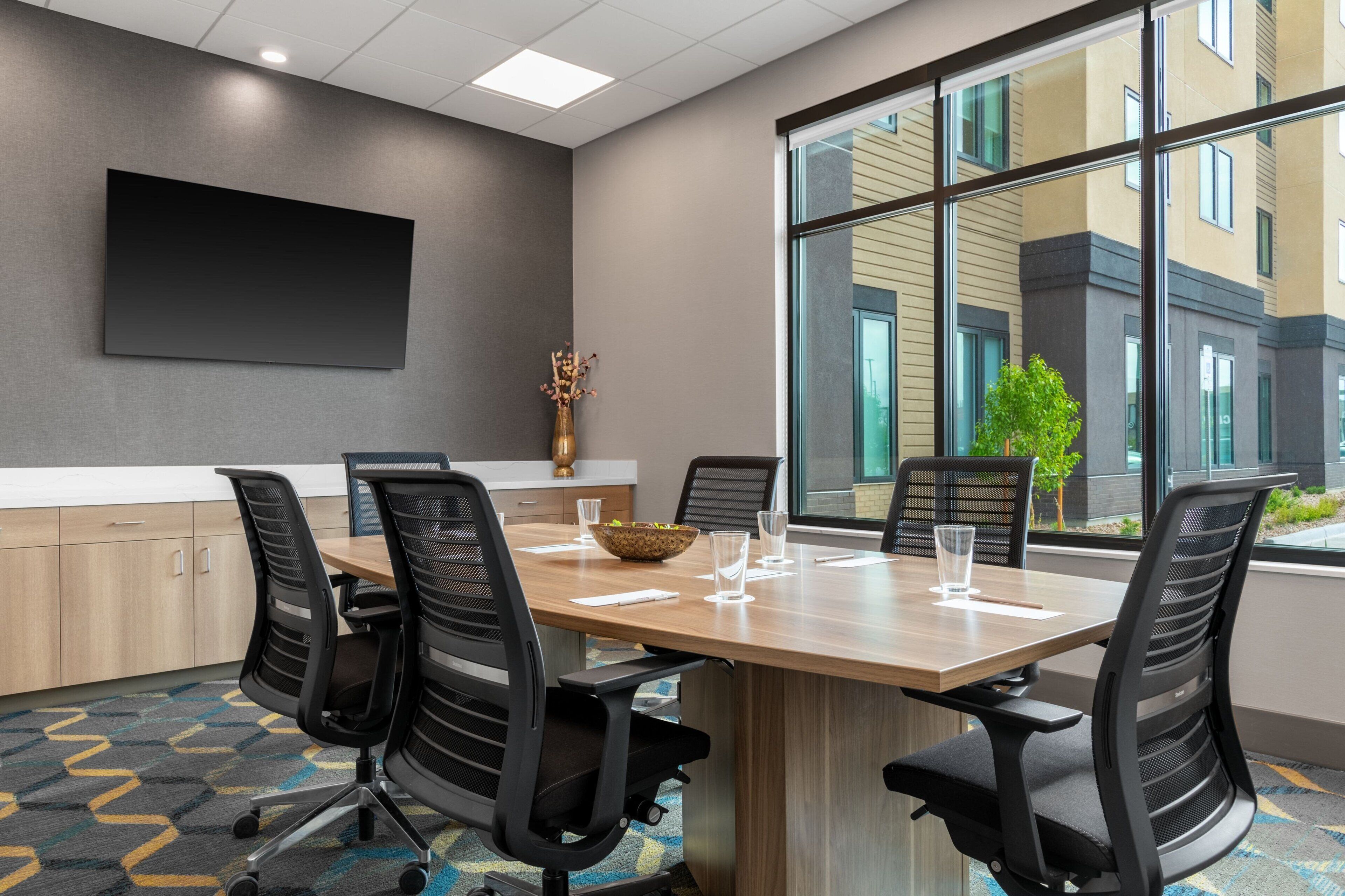 A conference room with a large table, six chairs, a flat-screen TV on the wall, and a window with a view of buildings.