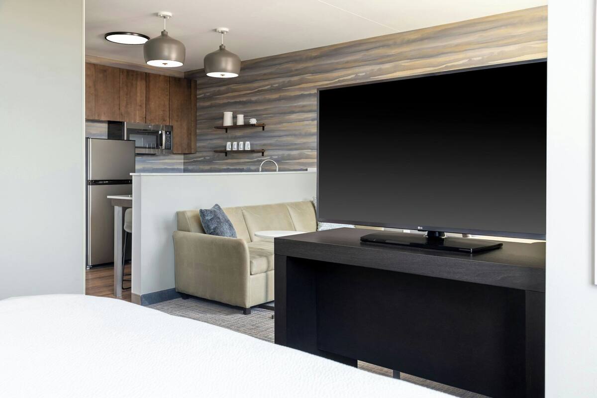 A modern hotel room with a kitchenette, a flat-screen TV, a sofa, and a bed.