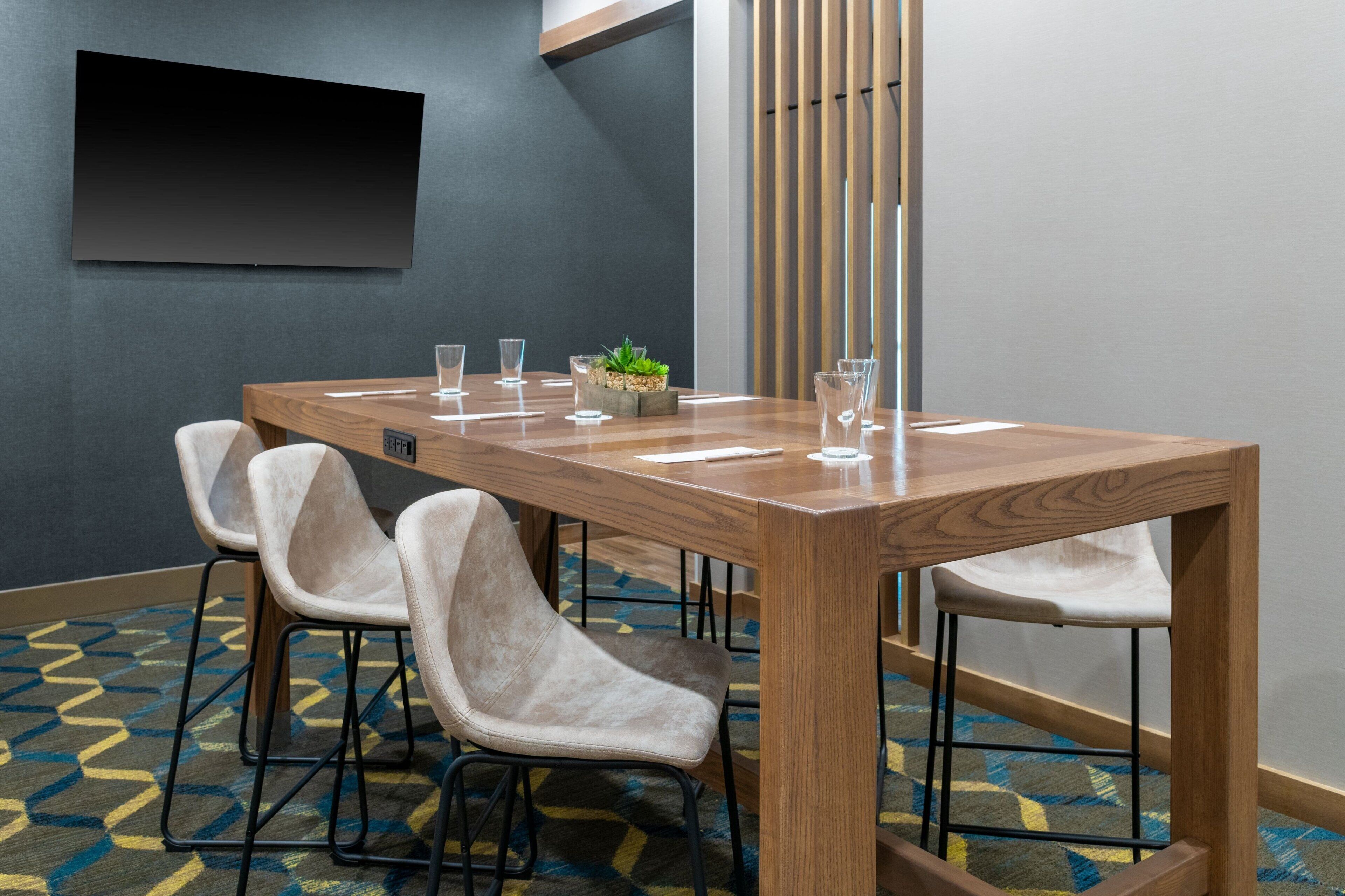 A conference room with a wooden table, four chairs, a small plant, and a flat-screen TV on the wall.