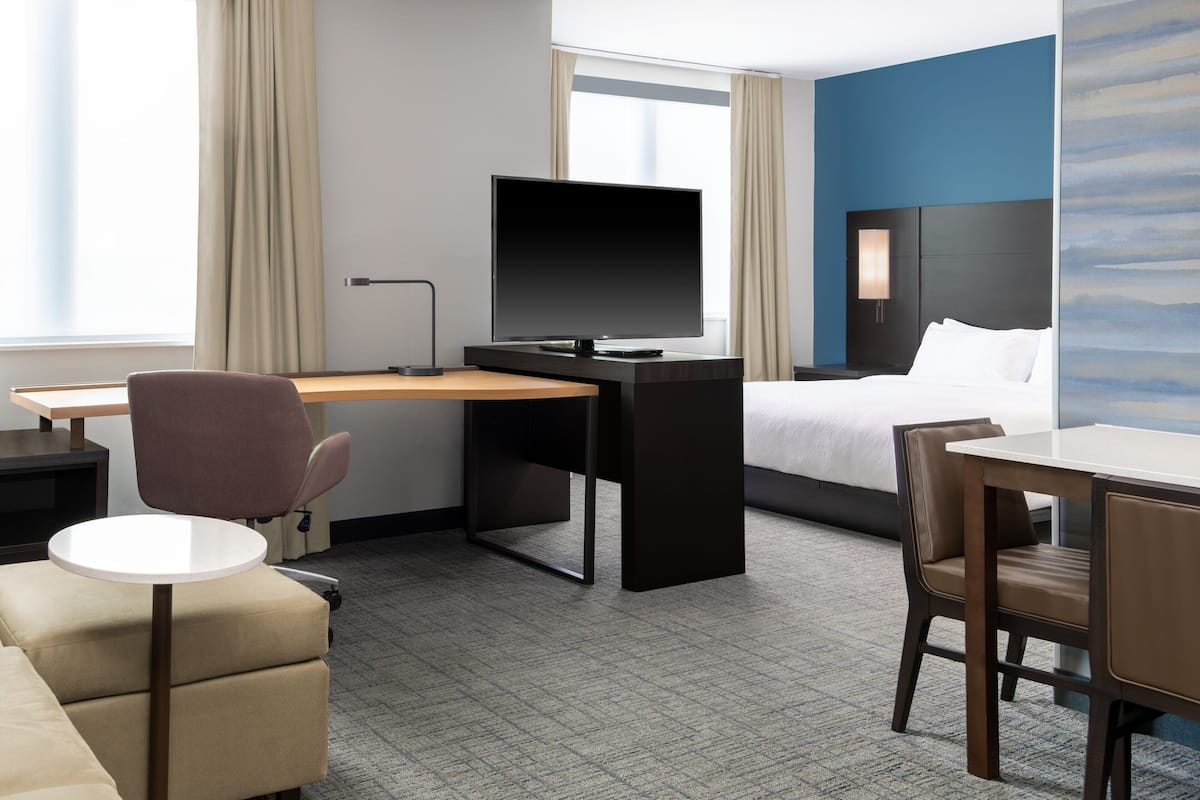A hotel room with a bed, a desk with a flat-screen TV, a chair, and a small table.