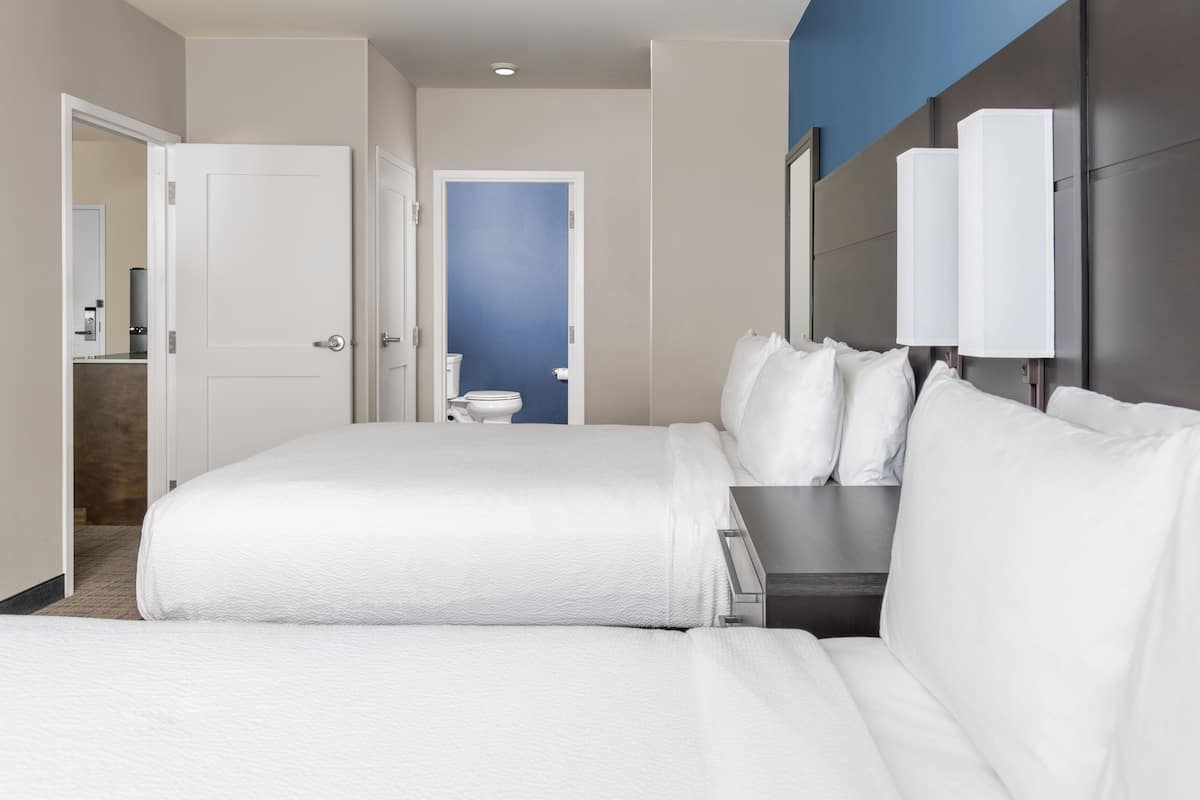 A hotel room with a large bed, a bedside table, a headboard with wall-mounted lamps, and a bathroom visible through an open door.