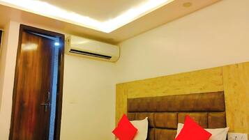 Habitación de hotel con una cama, ventilador de techo, unidad de aire acondicionado, cabecera de madera y mesita de noche con un teléfono y una planta.