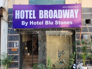 The entrance of Hotel Broadway by Hotel Blu Stones with a clear glass facade, security sign, and potted plants outside. - Hotel Broadway (New Delhi)