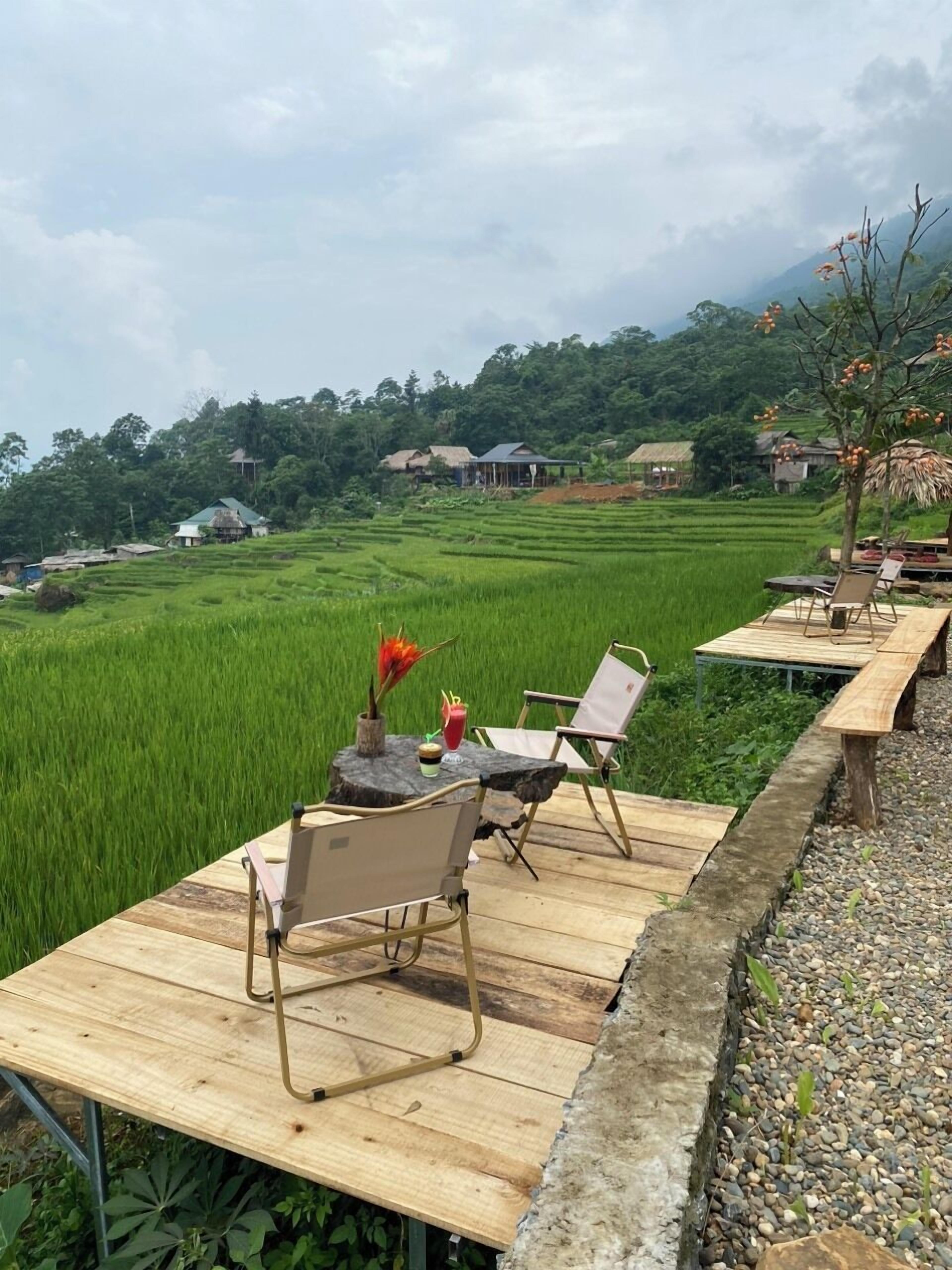 A serene outdoor seating area with wooden tables and chairs, overlooking lush green rice fields and traditional houses in the background.