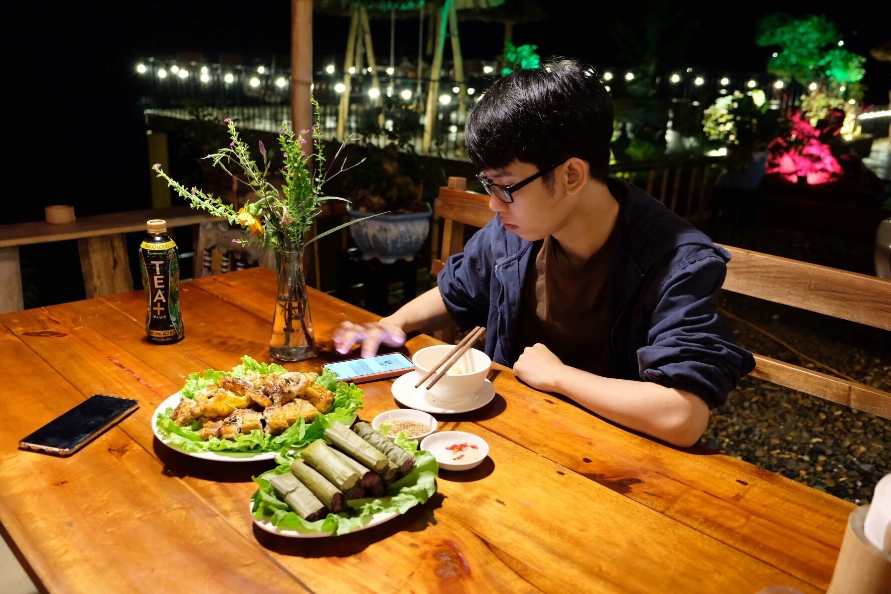 A person dining at an outdoor table with a variety of dishes, a bottle, and a smartphone.
