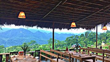 An outdoor seating area with wooden tables and benches, a thatched roof, and a view of mountains.