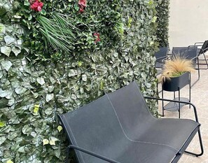 A black chair with a metal frame, positioned in front of a wall covered with green foliage and some red flowers. - MEDUSA BOUTIQUE (Athens)