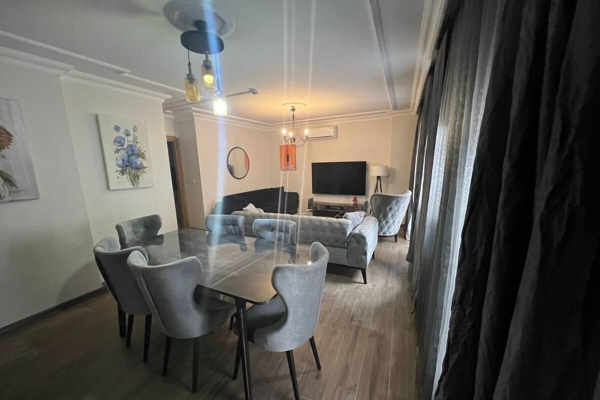 A modern living room with a glass-top dining table, grey upholstered sofas, a flat-screen TV, and a ceiling fan.