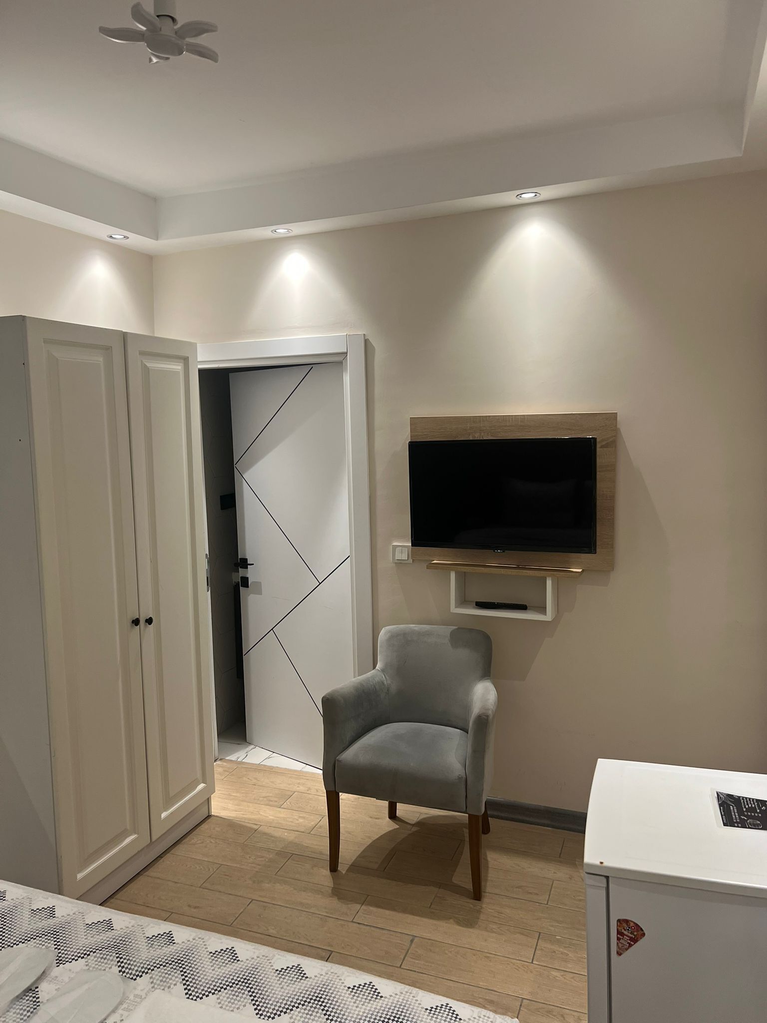 Una habitación con un sillón gris, un televisor montado en la pared, un armario blanco y una puerta que da a otra estancia.