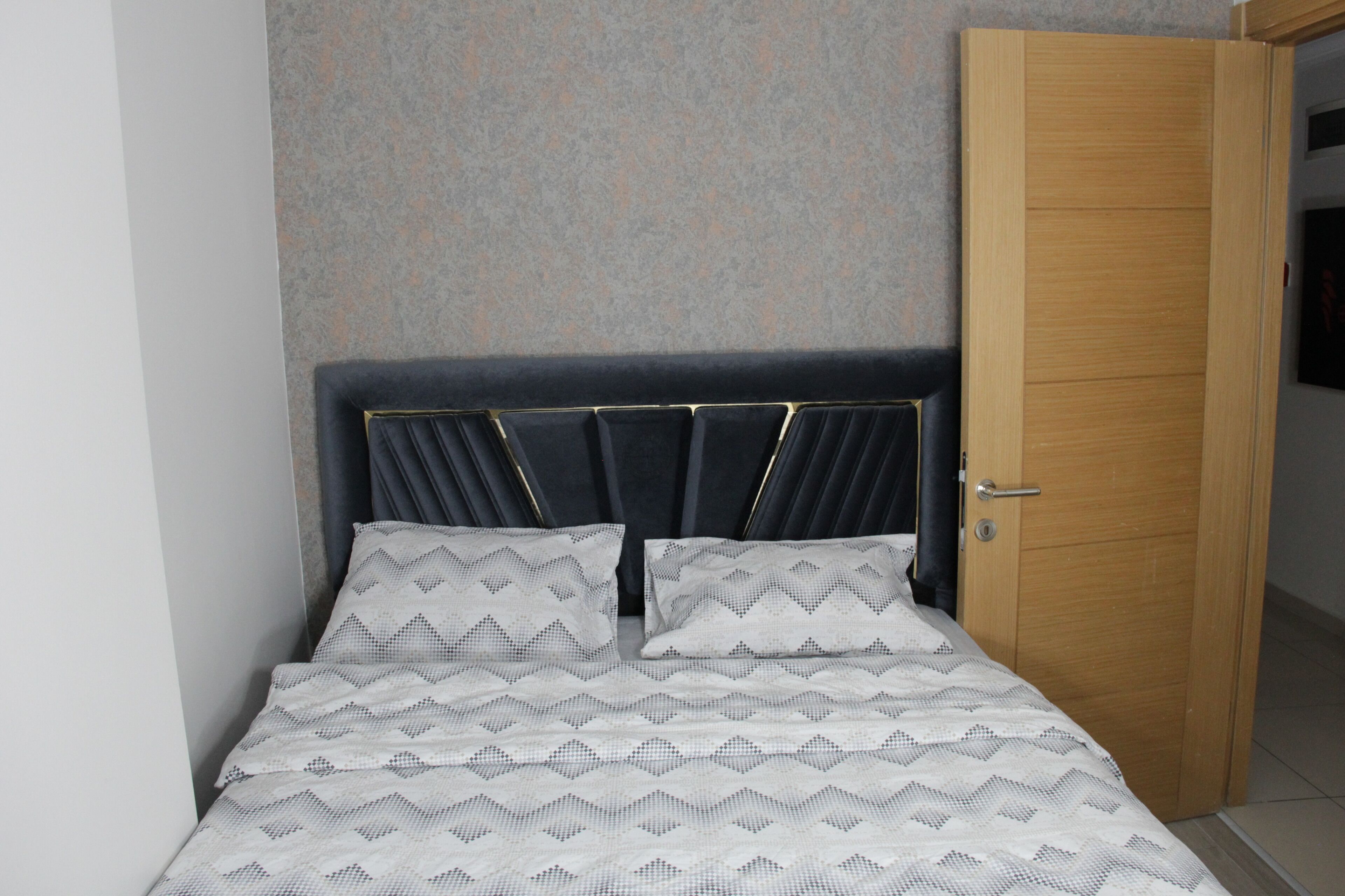 A bed with a patterned bedspread and two pillows, a wooden door, and a textured wall.