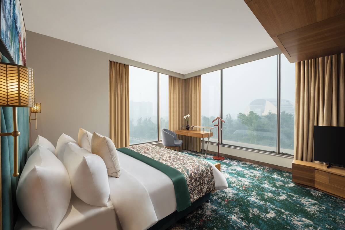 A hotel room with a large bed, a desk, a chair, a TV, and a view of the city through large windows.