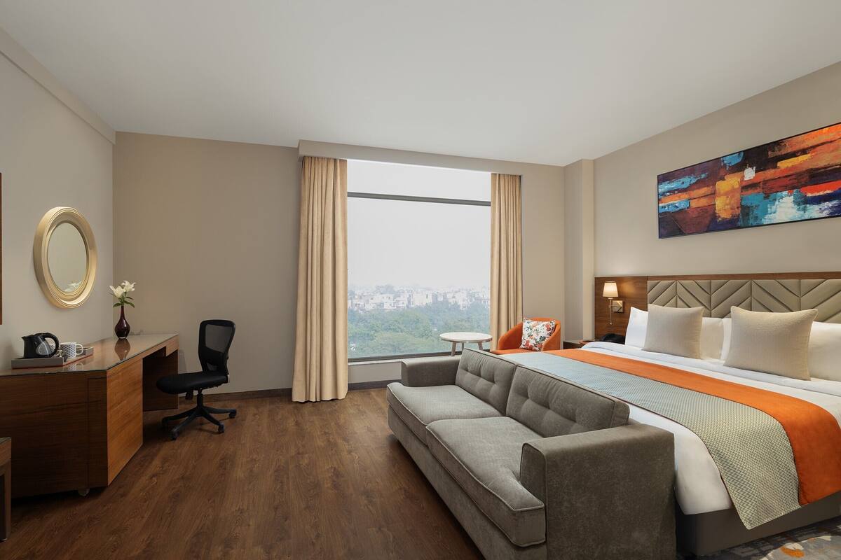 A hotel room with a bed, a sofa, a desk, a chair, a large window, and a painting on the wall.