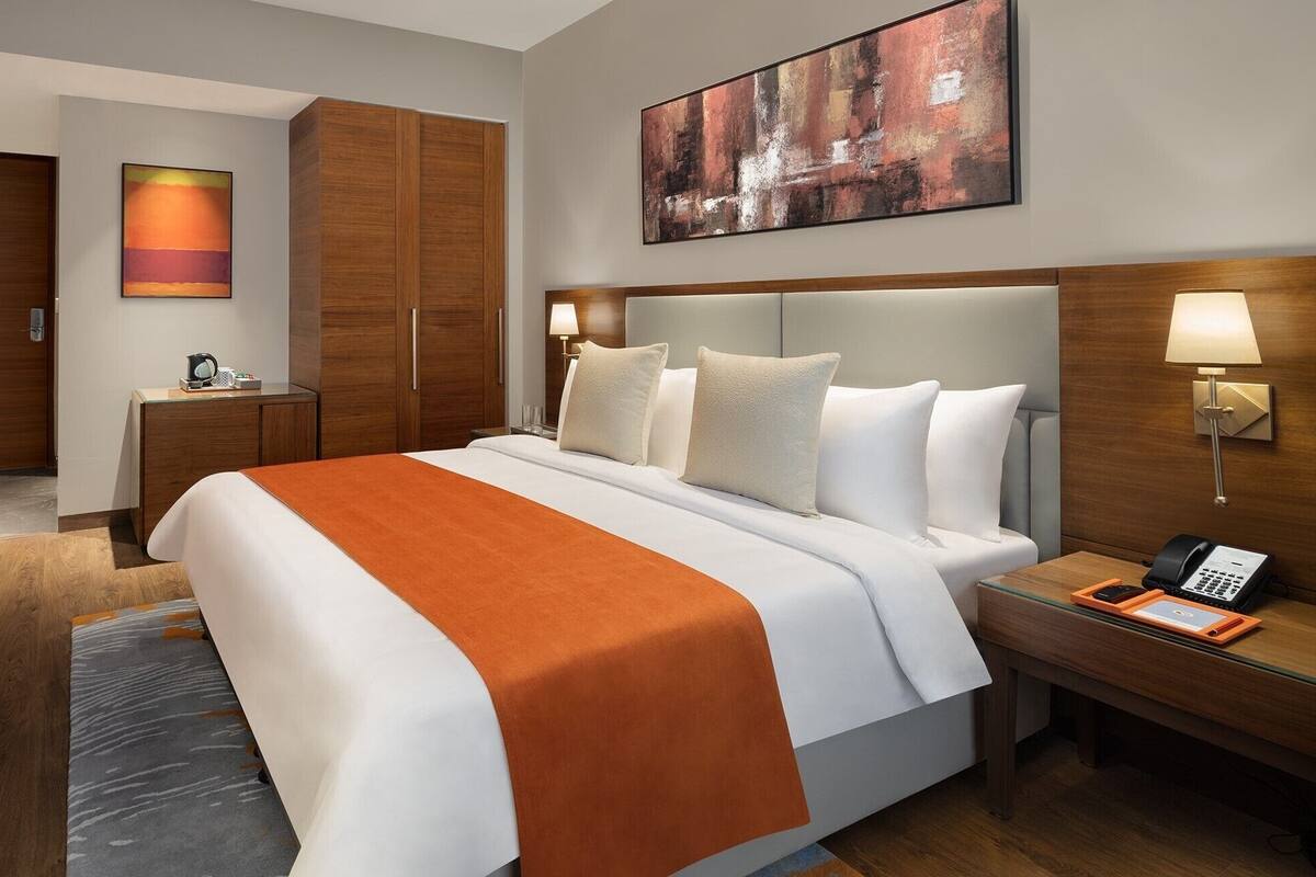 A hotel room with a large bed, a desk, a chair, a TV, a painting, and a lamp.