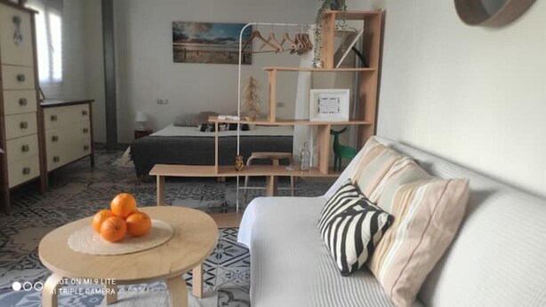 A modern living room with a sofa, a coffee table with oranges, a bed, a wooden bookshelf, and a painting on the wall.