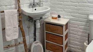 A bathroom with a white brick wall, a round mirror, a sink with a soap dispenser, a small wooden cabinet, and a towel hanging on a branch.