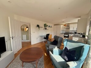Ein modernes Wohnzimmer mit einer Couch, einem Couchtisch und einem Fernseher. Im Hintergrund ist eine Küche mit Schränken und einem Herd zu sehen.