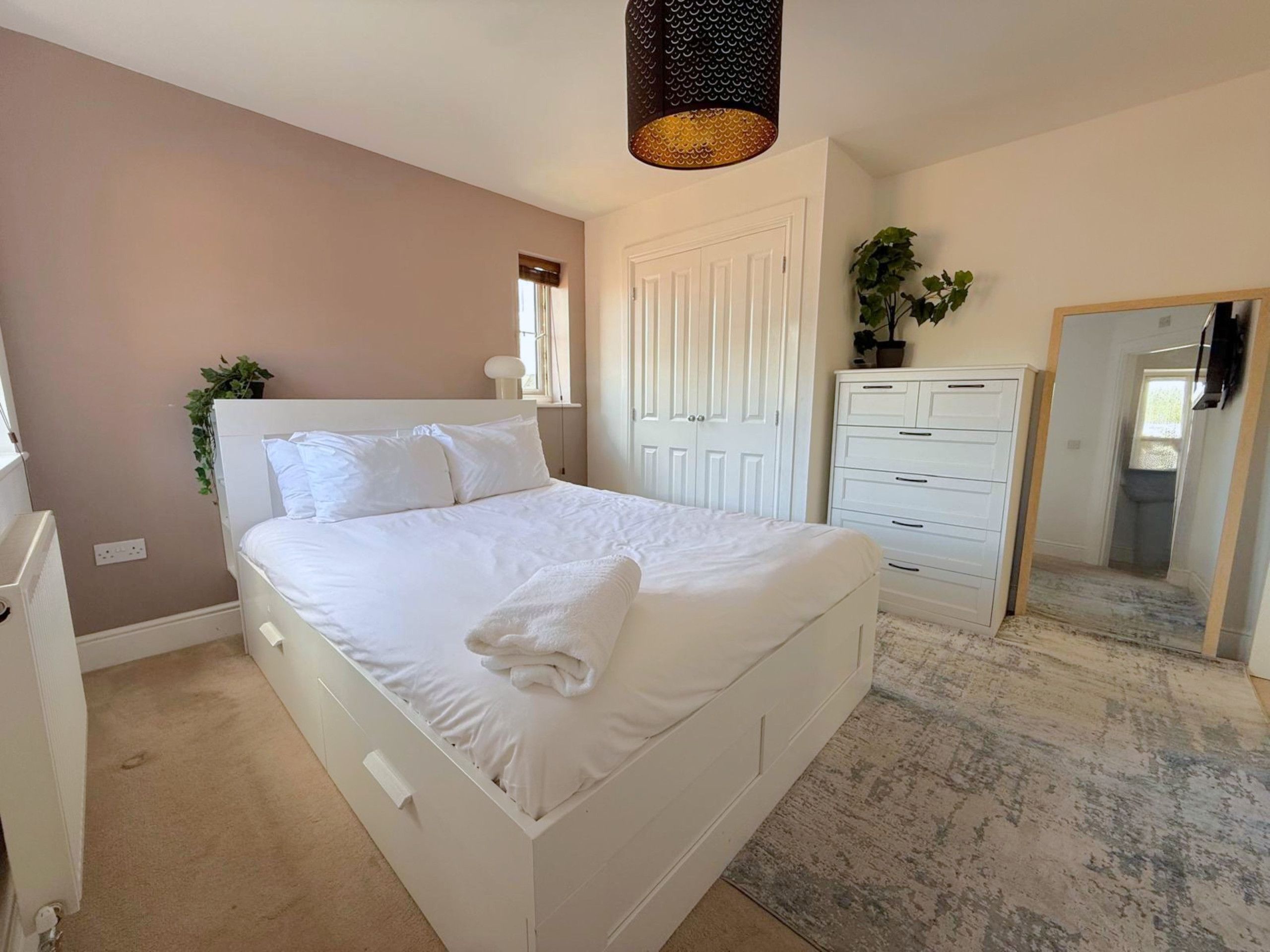 Un dormitorio con una cama blanca, un aparador, un espejo y una lámpara colgante.