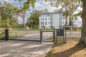 Une entrée fermée par un portail mène à un immeuble moderne, et un chemin pavé conduit à un espace de stationnement.