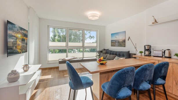 A modern kitchen with a dining area, wooden flooring, and a large window with blinds.