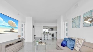 Ein modernes Wohnzimmer mit Flachbildfernseher, einem Glastisch, einer Couch mit gemusterten Kissen, einer Küche im Hintergrund und Bildern an den Wänden.