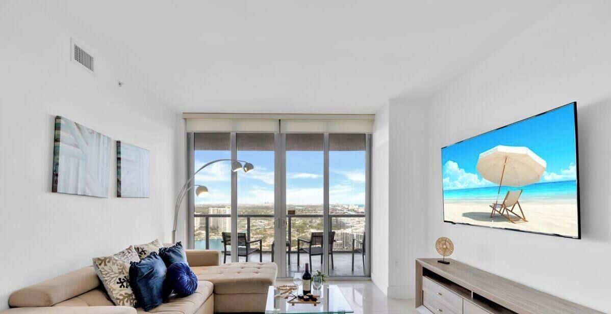 Een moderne woonkamer met een groot raam, een bank, een salontafel en een televisietoestel dat een strandscène toont.