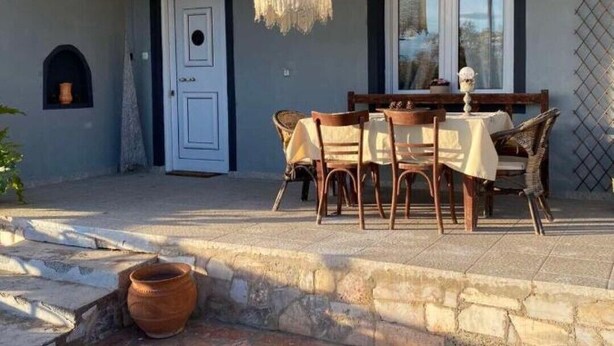 A covered outdoor dining area with a table and chairs, a hanging light fixture, and a stone wall.