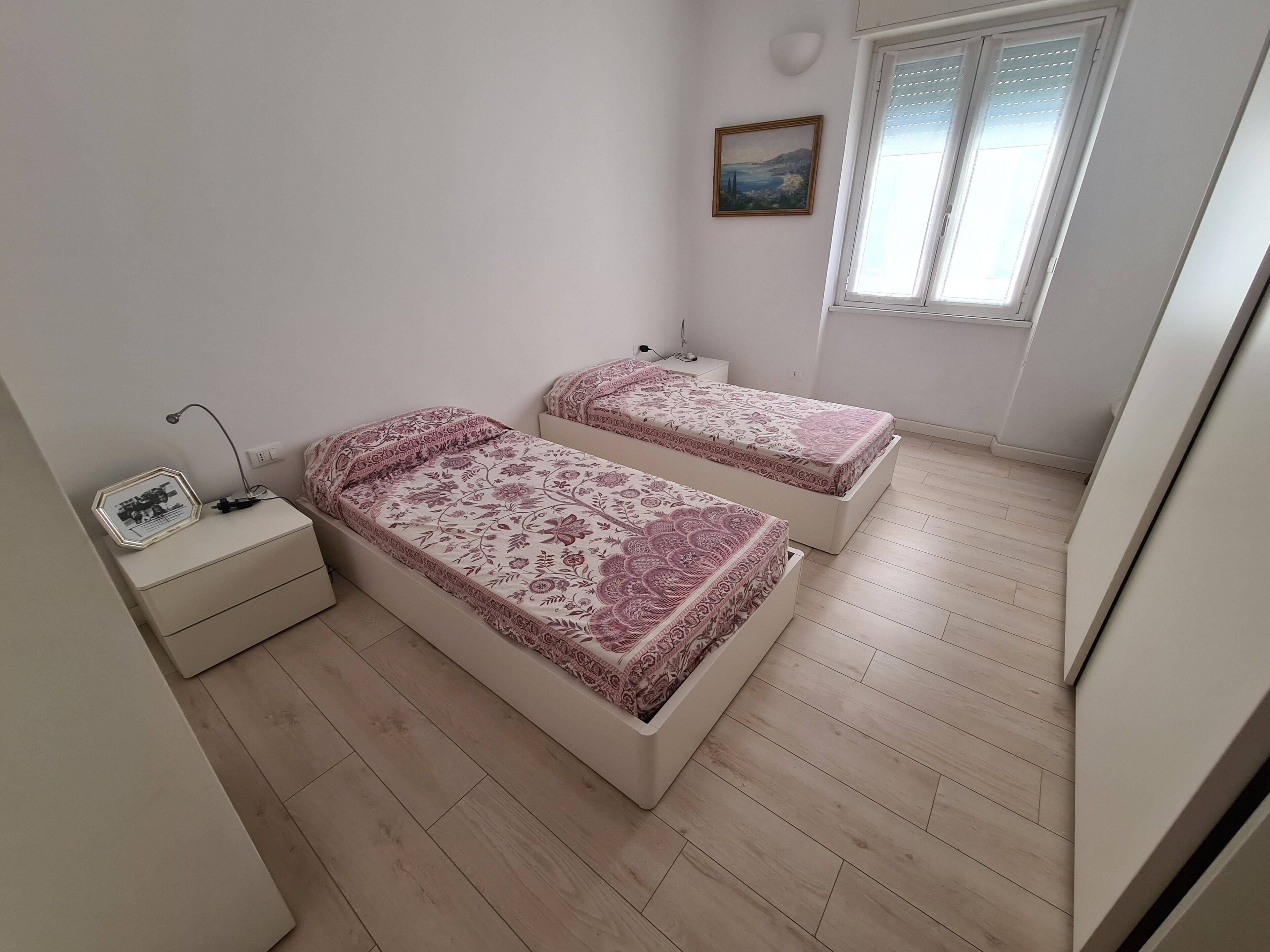 Une chambre avec deux lits, une table de chevet, une lampe fixée au mur, un tableau et une fenêtre avec des stores.