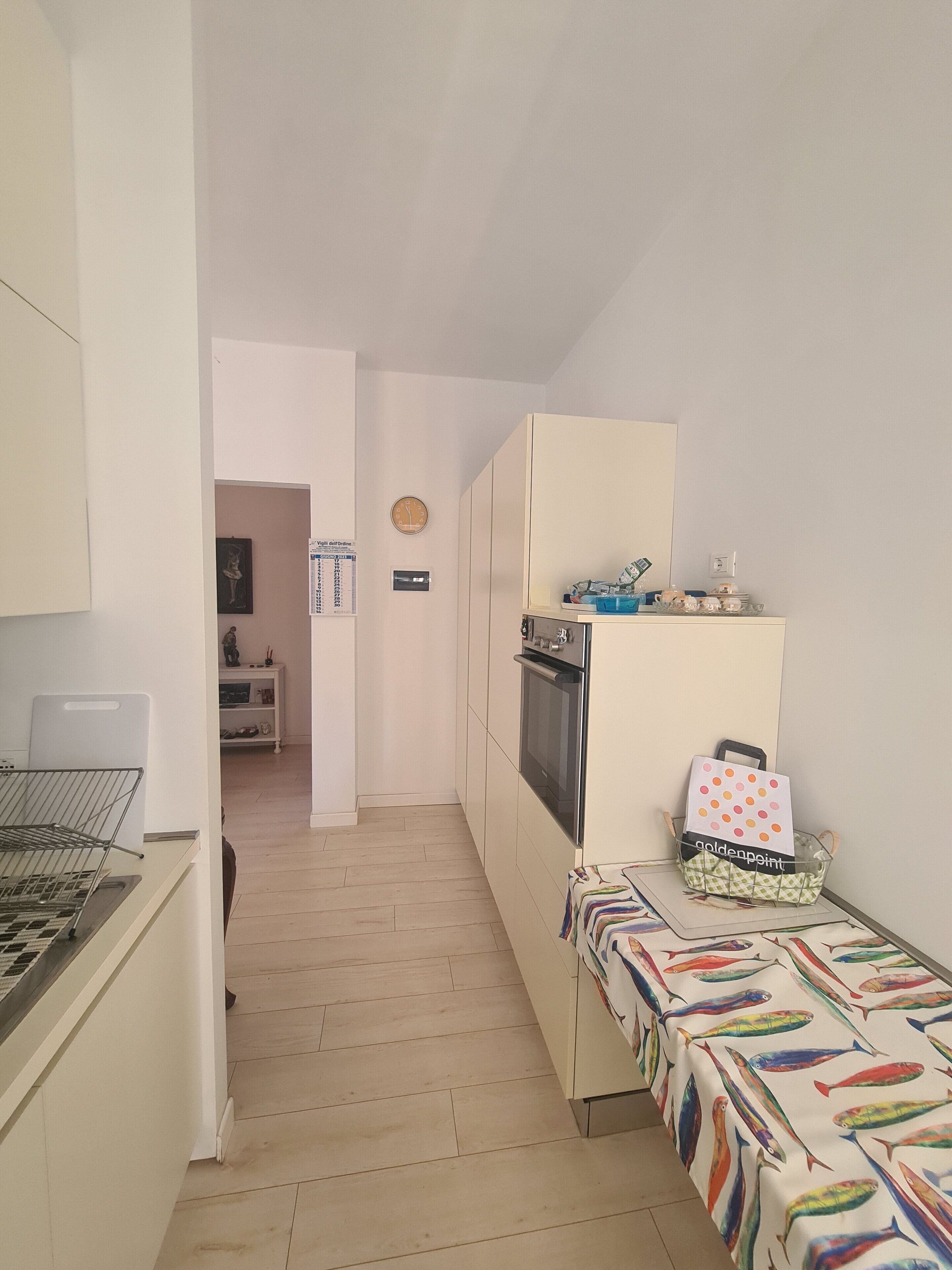 Cocina estrecha con estufa, horno y una pequeña mesa con mantel colorido. Se observa un armario con microondas y una cesta con tela de lunares. Un reloj es visible en la pared.