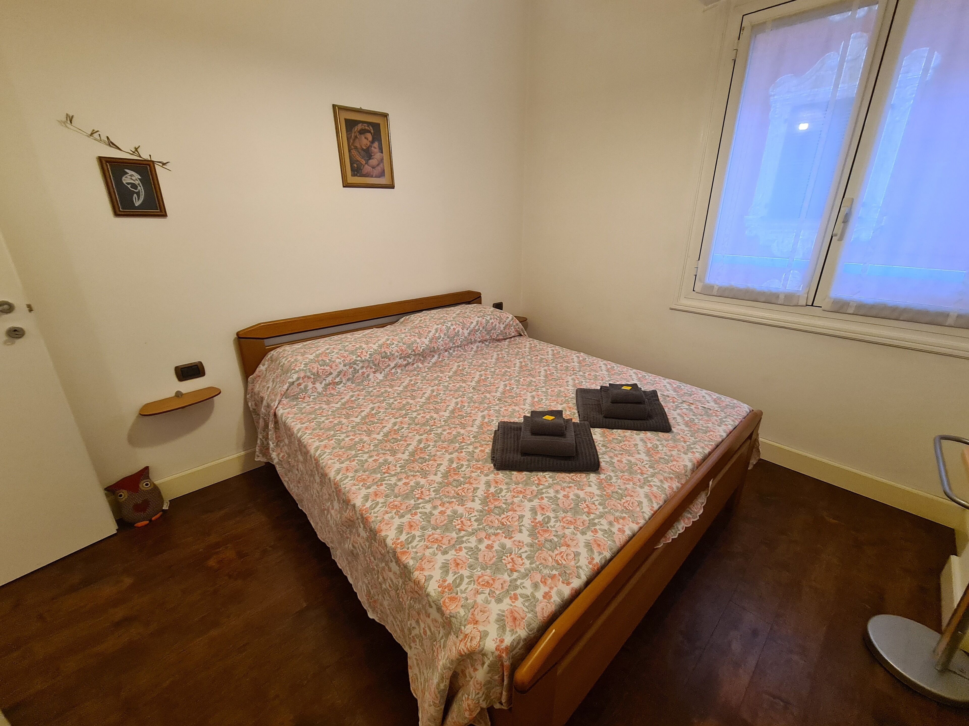 Une chambre avec un lit en bois, une couvre-lit à motifs floraux, deux serviettes, une petite étagère et deux tableaux encadrés accrochés au mur.