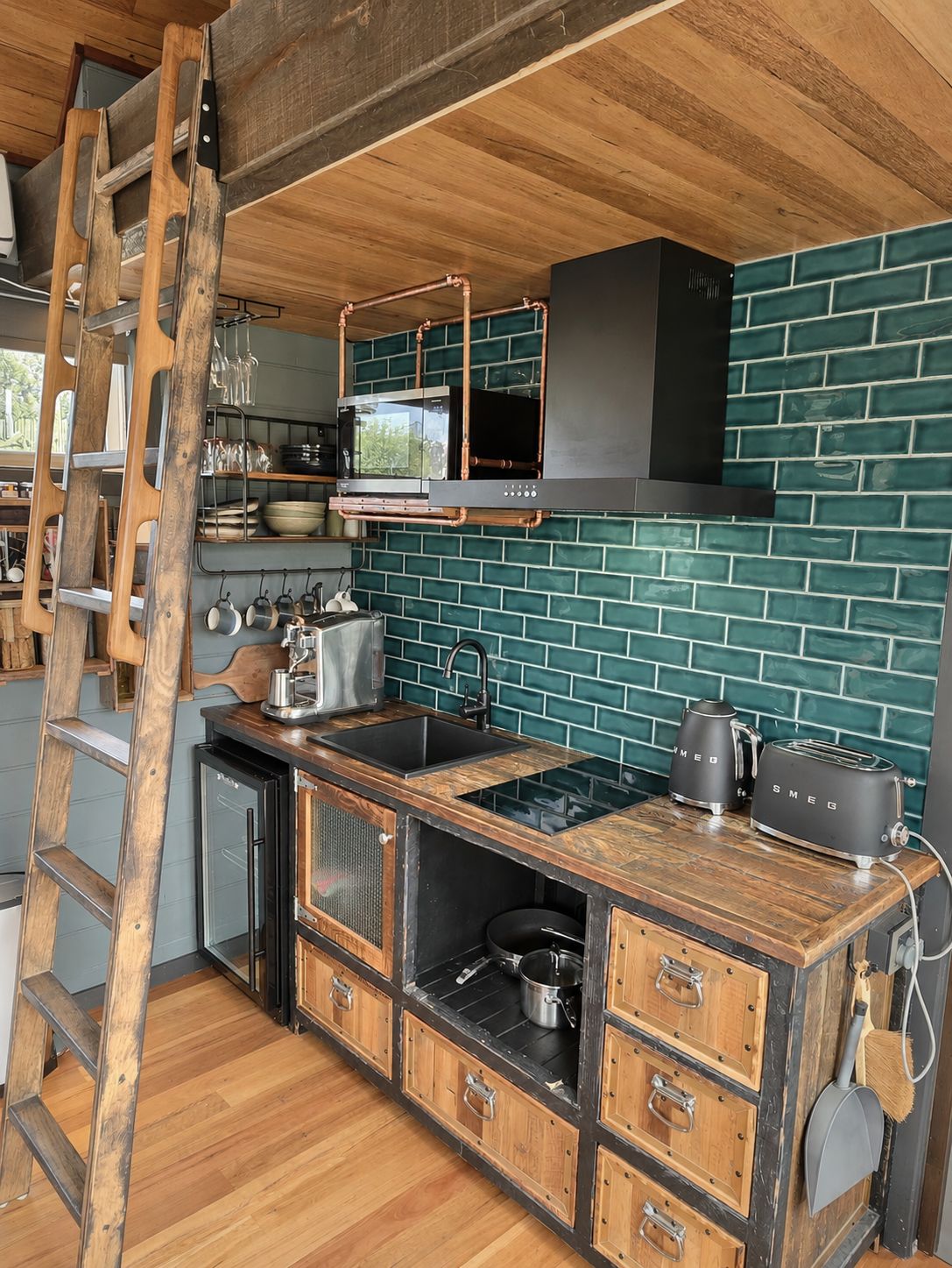 Une cuisine avec une échelle en bois, un poêle noir et un pare-feu vert en carreaux.