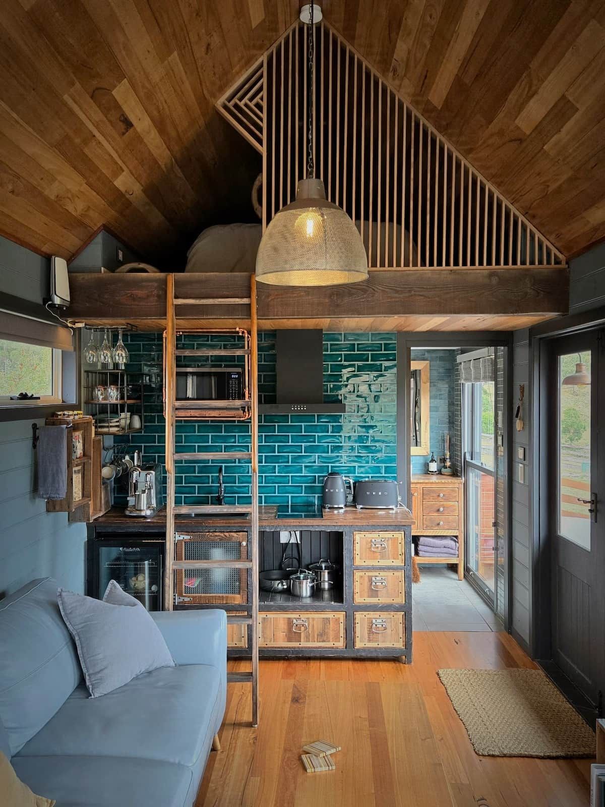 Ein modernes Interieur mit Holzboden, einer hängenden Leuchte, einem blauen Sofa und einer Küchenzeile mit Backsteinwand.