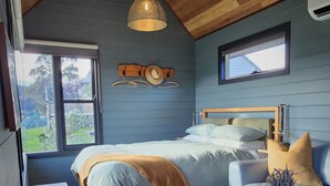 Een gezellige slaapkamer met een houten plafond, een bed met een blauw hoofdeinde, een houten bankje, een kleine tafel en een raam met uitzicht op groen.