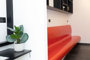 A reception area with a potted plant on a black shelf, a red bench, and a wall-mounted light switch. - Citizen suites (Sliema)