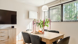 Un comedor con una mesa de madera, sillas negras, un jarrón con flores y un televisor sobre un mueble.