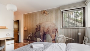Un dormitorio con una gran escultura de elefante, una cama con sábanas blancas, un escritorio de madera y una ventana con cortinas.