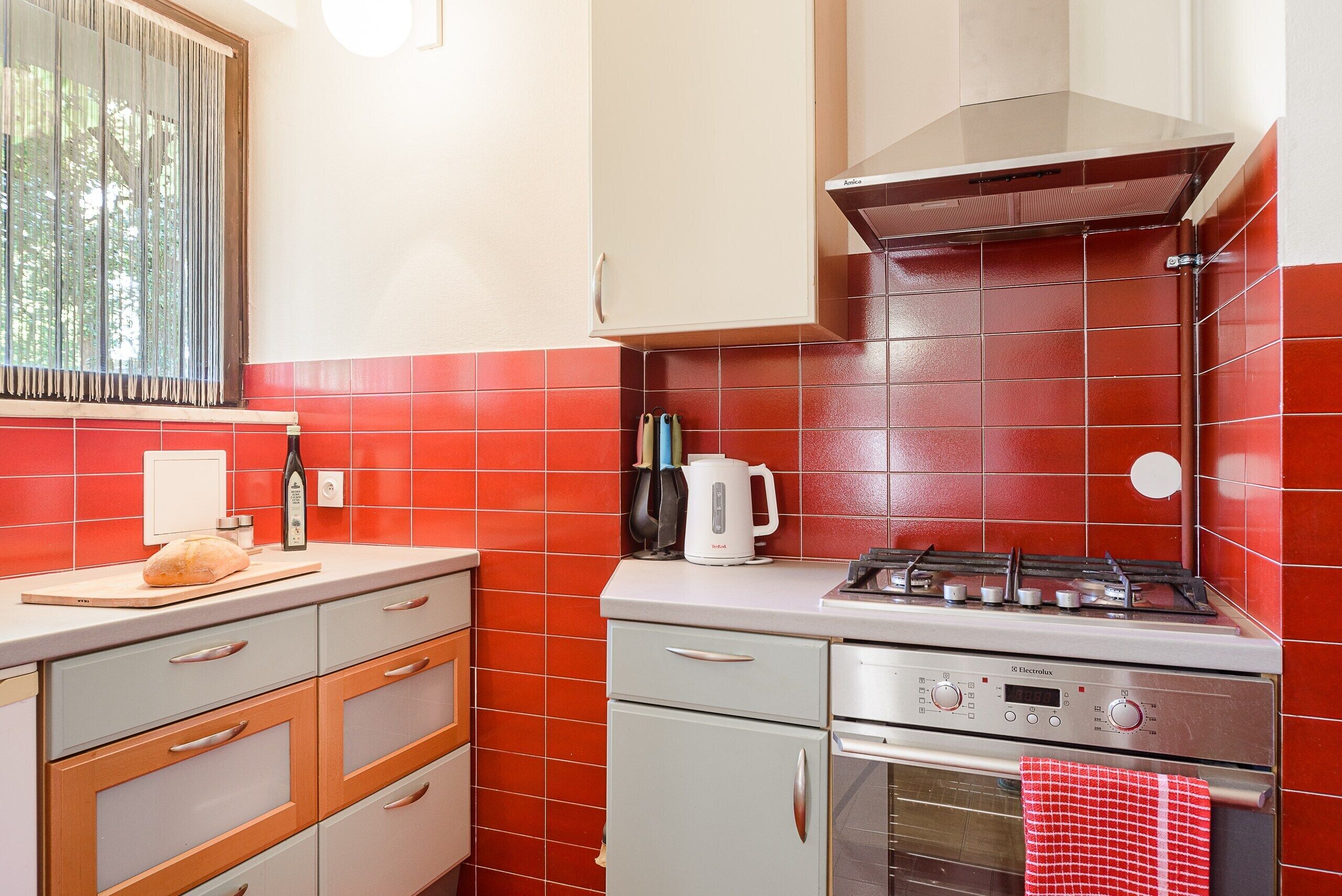 A kitchen with red tiles, a gas stove, a built-in oven, and a window with curtains.