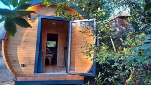 Una cabaña de madera con una puerta grande abierta, un interior visible con una cama y una chimenea, rodeada de follaje denso.