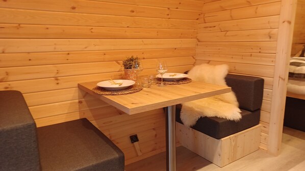 A wooden cabin interior with a dining table set for two, a small potted plant, and a white furry chair.