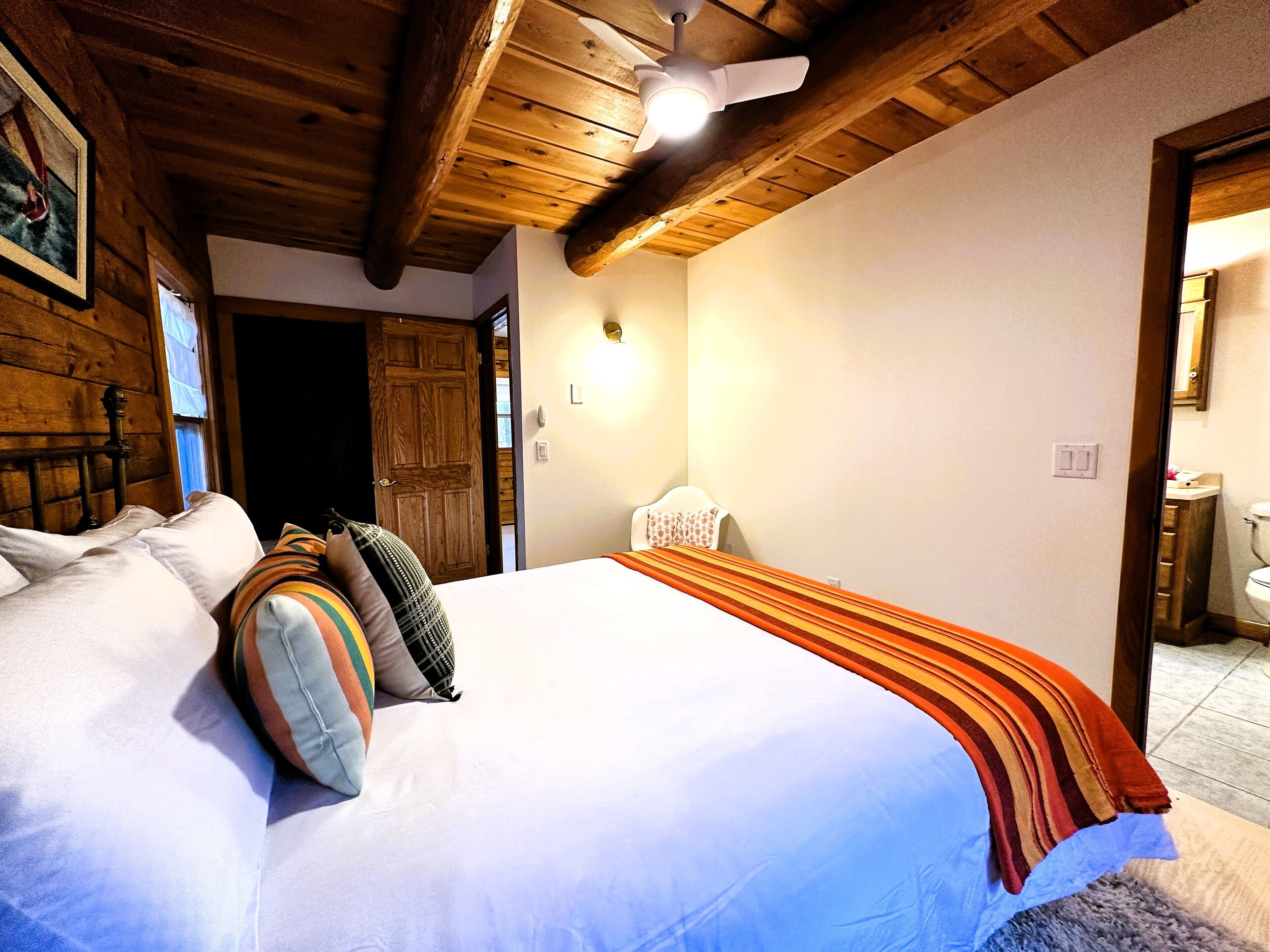 Un dormitorio con techo de madera, cama con ropa de cama blanca y manta a rayas.