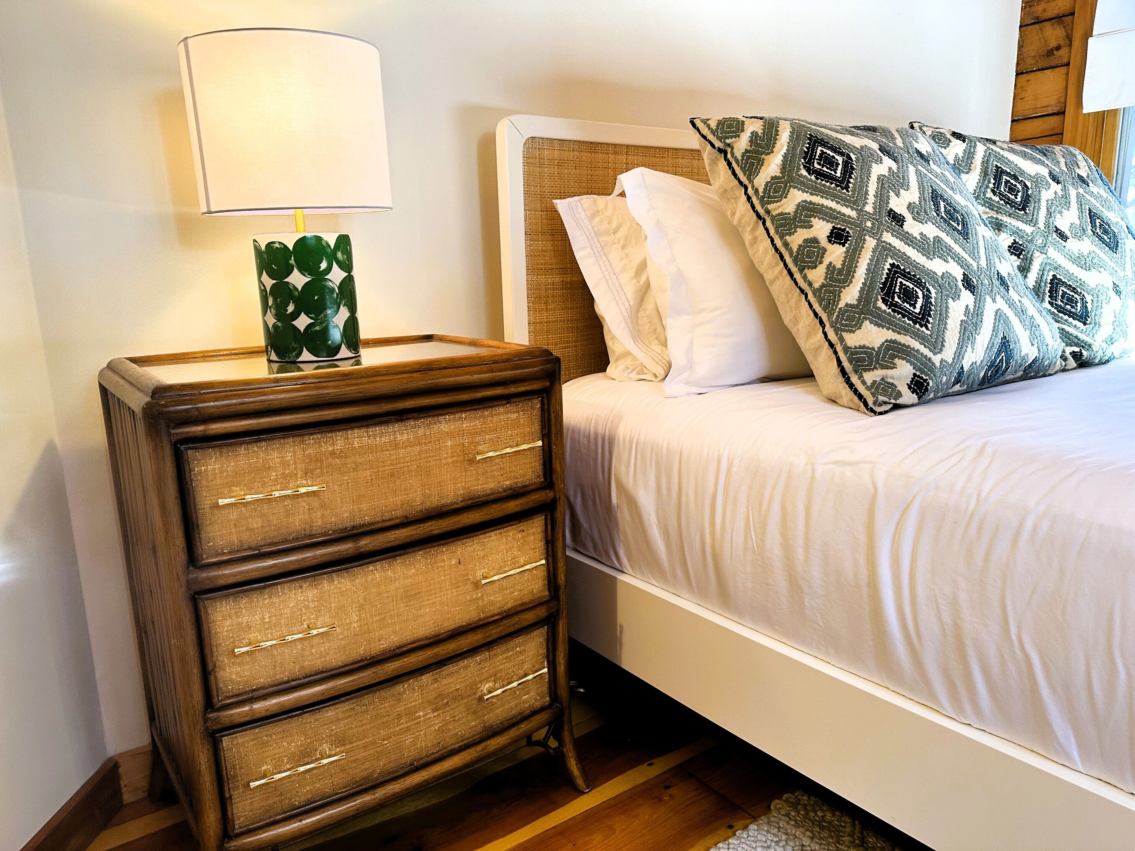 A bed with white bedding and a patterned pillow, a wooden nightstand with drawers, and a lamp with a green vase.