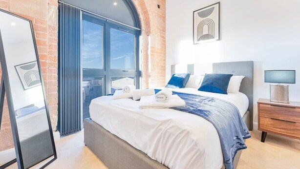 Une chambre d’hôtel moderne avec un grand lit, un miroir, une table de chevet et une vue sur l’extérieur, visible à travers une porte vitrée.