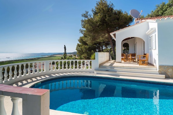 A pool with a white balustrade, a covered patio with outdoor furniture, and a view of the sea.